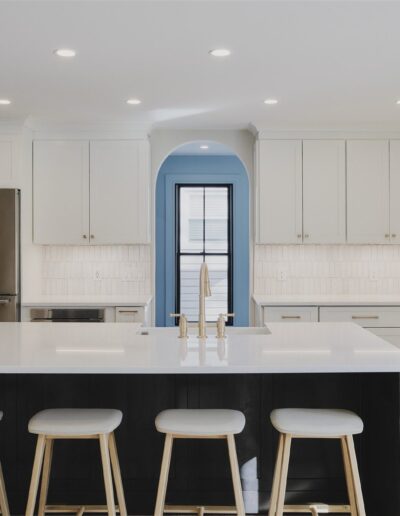 Modern kitchen with white cabinets, stainless steel appliances, large island with white countertop, four wooden stools, and a central archway leading to another room.