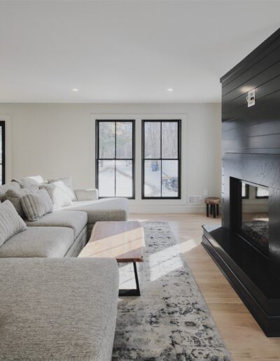 Modern living room with a large gray sectional sofa, patterned rug, wooden coffee table, and a black shiplap fireplace. Two large windows let in natural light.