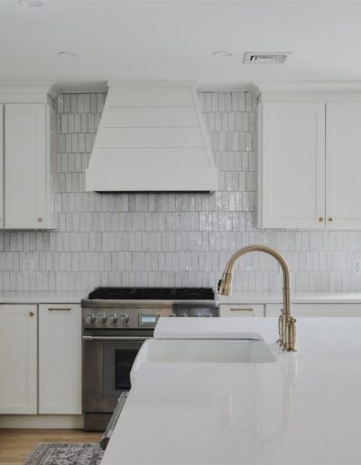Modern kitchen with white cabinets, white tile backsplash, stainless steel stove, gold faucet, and large white island next to glass doors.