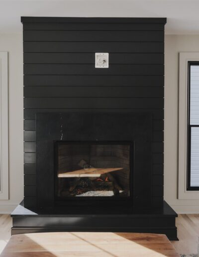 A modern black fireplace with a wood mantel is centered between two large windows in a bright, minimalist living room.