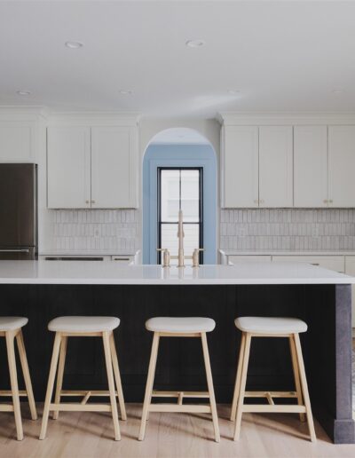 Modern kitchen with white cabinets, stainless steel appliances, large island with white countertop, and four wooden stools. Light wood flooring and neutral tile backsplash are visible.