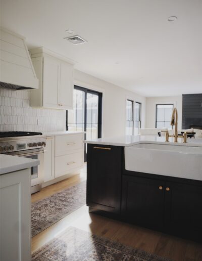 Modern kitchen with white cabinetry, a large farmhouse sink on a dark island, brass fixtures, stainless steel appliances, and patterned rugs on a wood floor.