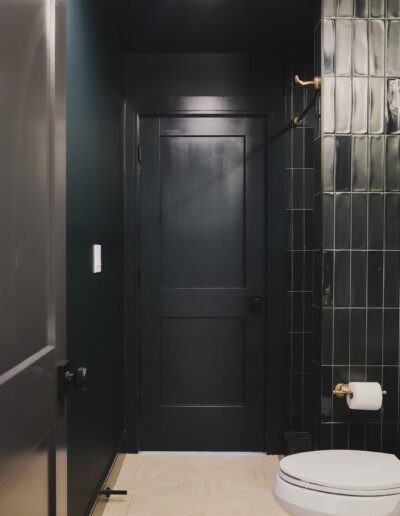 A modern bathroom with dark walls, black door, glossy black tiled shower, white toilet, and a white sink with brass fixtures.