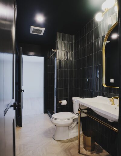 Modern bathroom with black tiled walls, a white toilet, a white sink with gold fixtures, a gold-framed mirror, and light wood hexagonal floor tiles.