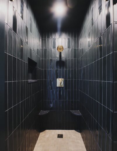 A modern shower with dark glossy tile walls, a built-in niche, two small corner shelves, a square drain, and an overhead showerhead under a ceiling light.
