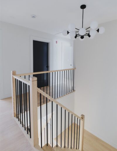 A modern staircase landing with light wood flooring, black metal railings, white walls, black door, and a contemporary black and white chandelier.