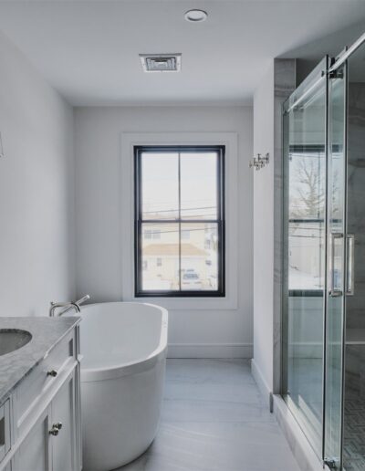 Modern bathroom with a marble countertop, white vanity, freestanding bathtub, glass shower enclosure, and a large window letting in natural light.
