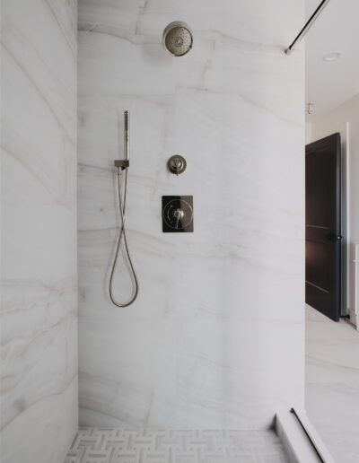 Modern shower with white marble walls, a rainfall showerhead, a handheld showerhead, and metal controls, next to a glass panel and mosaic tile floor.