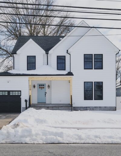 Two-story white house with black trim and a one-car garage, surrounded by snow, on a residential street with neighboring homes.