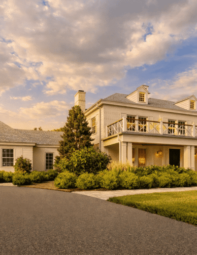 Two-story suburban house with a wraparound balcony, large windows, neatly trimmed shrubs, and a paved driveway under a partly cloudy sky at sunset.